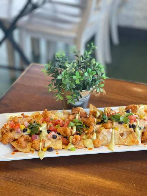 A platter of nachos topped with fried chicken, tomatoes, black beans, and drizzled with sauce, set on a wooden table.