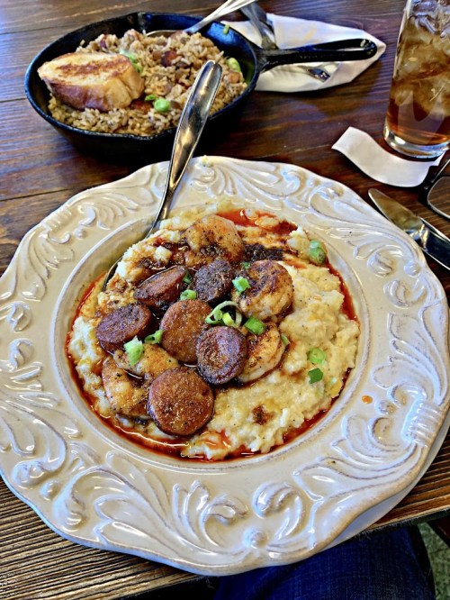A plate of creamy grits topped with sliced sausage and green onions, with a side of rice and iced tea in the background.