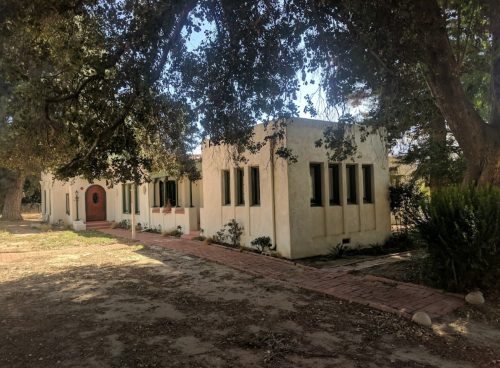 A quaint, stucco building surrounded by trees, with a pathway leading to a round entrance.