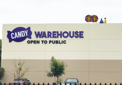 A building with a sign reading "Candy Warehouse Open to Public," featuring two people on the roof with colorful balloons.