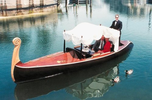 A gondola with a canopy floats on water, featuring two passengers and a gondolier in formal attire. Ducks swim nearby.