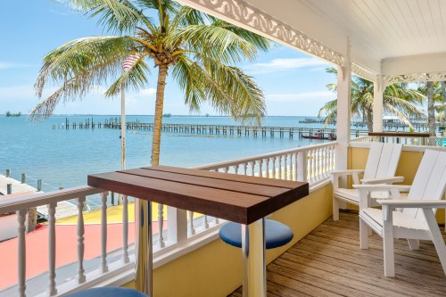 A scenic view from a porch with white chairs, overlooking a calm sea and palm trees, with a pier in the background.