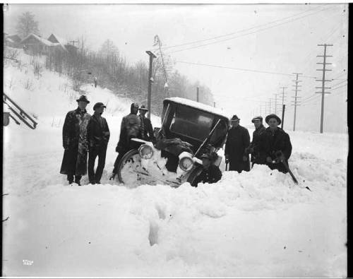 A group of men stands beside a vintage car stuck in deep snow, with power lines and snowy hills in the background.