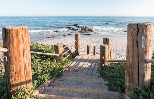 Wooden steps lead down to a sandy beach with gentle waves and rocky outcrops under a clear blue sky.