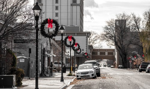 A festive street scene with wreaths and red bows on lamp posts, lined with parked cars and buildings in the background.