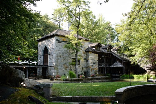 A stone building surrounded by trees, featuring a wooden structure and a small pond in the foreground.