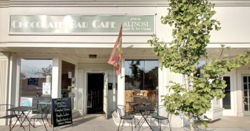Exterior of Chocolate Bar Café featuring a sign, outdoor seating, and a flag, with a chalkboard menu displayed.