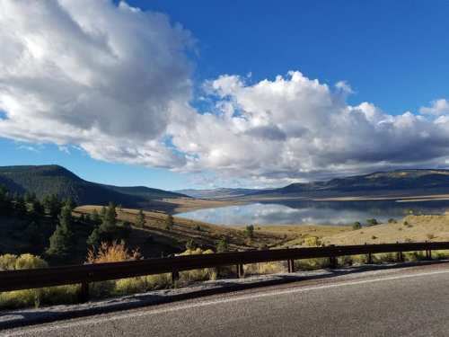 Scenic view of a lake surrounded by hills and trees, with fluffy clouds in a blue sky reflecting on the water.