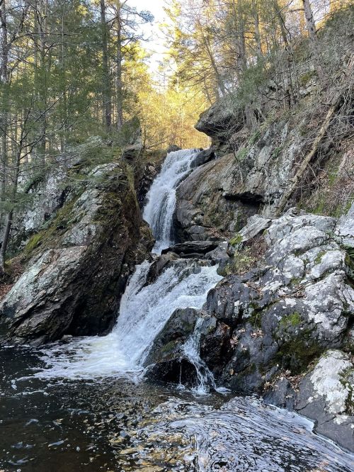 A serene waterfall cascading down rocky terrain, surrounded by trees and sunlight filtering through the forest.