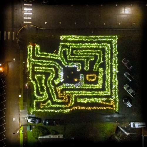 Aerial view of a maze-shaped light display in a parking lot, surrounded by parked cars at night.
