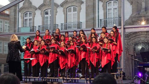 A choir of young women in red capes performs on stage, led by a conductor, with a historic building in the background.