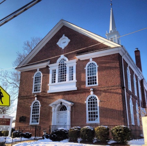 Historic brick church with a white steeple, surrounded by snow and greenery, under a clear blue sky.