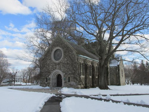 A stone church surrounded by snow, with a large tree and a blue sky filled with clouds in the background.