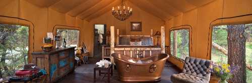 Luxurious glamping tent interior with a copper bathtub, chandelier, and cozy furnishings surrounded by nature.