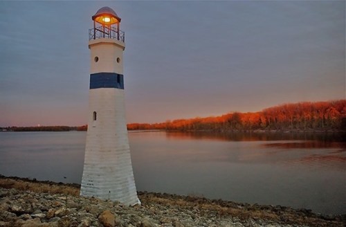 A white and blue lighthouse stands by a calm river at dusk, with trees reflecting warm colors in the background.