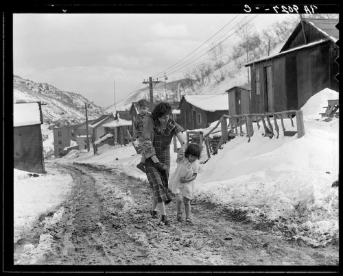 A woman carries a child while holding another child's hand, walking down a snowy road lined with wooden houses.
