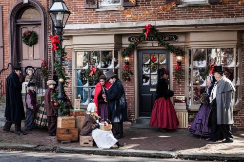 A festive street scene with people in Victorian attire, shopping and celebrating the holidays outside a decorated store.