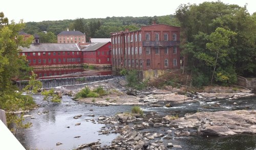 Historic red brick buildings beside a river with a small waterfall and rocky shoreline, surrounded by greenery.