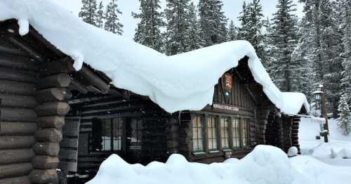 A snow-covered log cabin surrounded by tall trees in a winter landscape. Snow blankets the roof and ground.