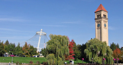 A park scene featuring a tall clock tower and a large, modern structure against a clear blue sky and colorful trees.