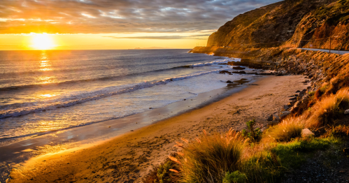 Sunset over a serene beach with gentle waves, rocky cliffs, and a winding road along the coastline.