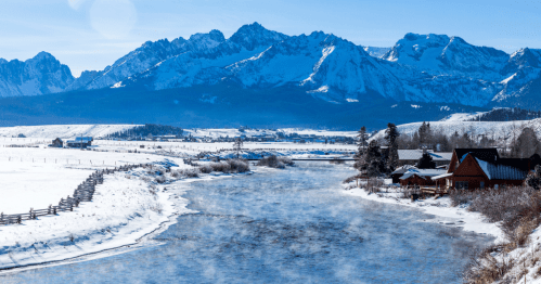 Snow-covered mountains rise above a winding river, with a rustic cabin and trees along the snowy banks.