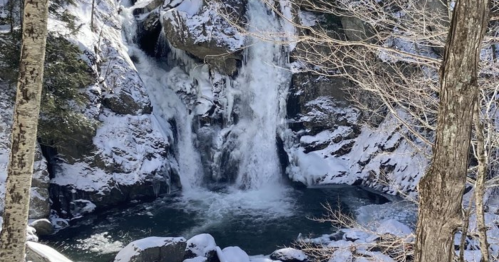 A snowy waterfall cascading into a pool, surrounded by rocky cliffs and bare trees.