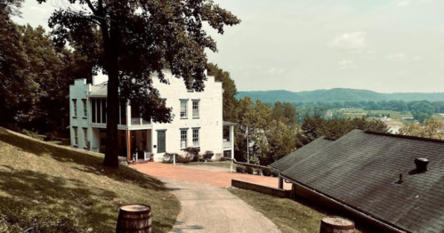 A white building on a hillside with a winding path, surrounded by trees and a scenic view of rolling hills.