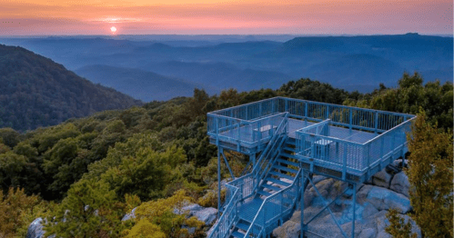 A viewing platform on a mountain at sunset, surrounded by lush green trees and distant blue hills.