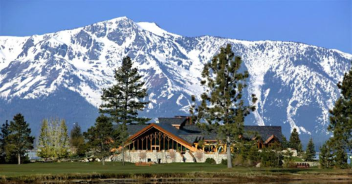 A scenic view of a rustic lodge surrounded by trees, with snow-capped mountains in the background under a clear blue sky.