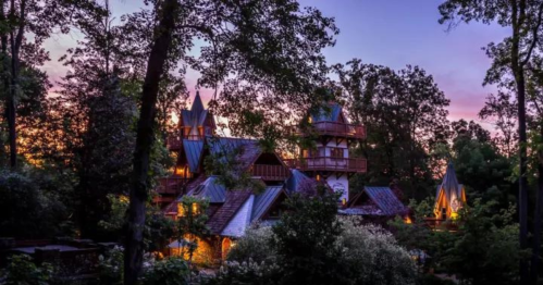 A whimsical, fairy-tale style house surrounded by trees at dusk, with warm lights glowing from its windows.