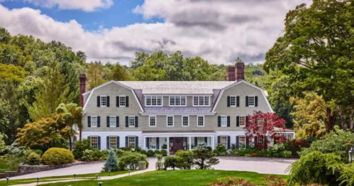 A large, elegant green house surrounded by lush trees and colorful landscaping under a partly cloudy sky.