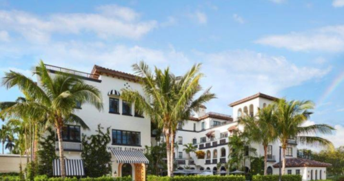 A charming hotel with palm trees, featuring a white facade and striped awnings under a blue sky with a rainbow.