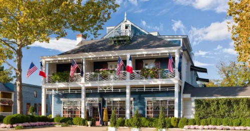 A charming blue house with a large porch, adorned with flags and surrounded by greenery and flowers under a clear sky.