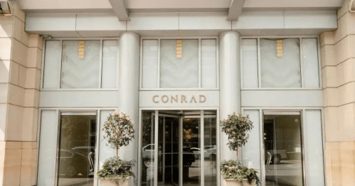 Entrance of the Conrad hotel, featuring large glass doors and decorative potted plants on either side.