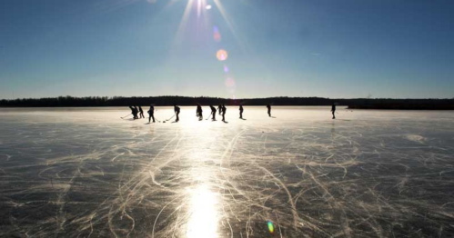 Silhouettes of people skating on a frozen lake under a bright sun, with visible ice patterns and a clear blue sky.