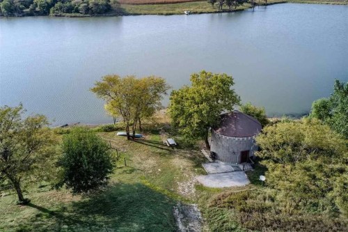 Aerial view of a small round building by a calm lake, surrounded by trees and grassy areas.