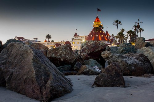 View of a historic hotel with a red roof, surrounded by palm trees and large rocks on a sandy beach at dusk.
