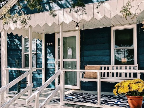 A blue house with a porch, featuring a bench, potted flowers, and a patterned rug.
