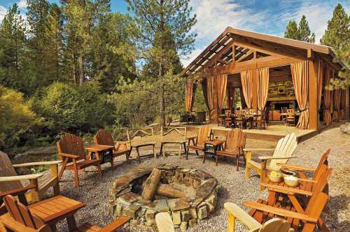 A cozy outdoor seating area with wooden chairs around a fire pit, near a rustic cabin surrounded by trees.