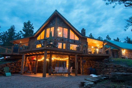 A cozy mountain cabin with large windows, a deck adorned with string lights, and a stone exterior, set against a twilight sky.