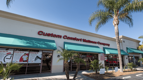 Exterior of Custom Comfort Mattress Company store with palm trees and blue sky. Sleep tips displayed in windows.