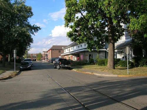 A quiet street scene with parked cars, trees, and buildings under a partly cloudy sky.