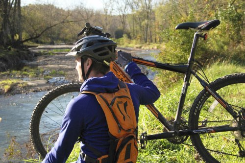 A cyclist in a helmet carries a mountain bike by a stream, surrounded by trees and greenery.