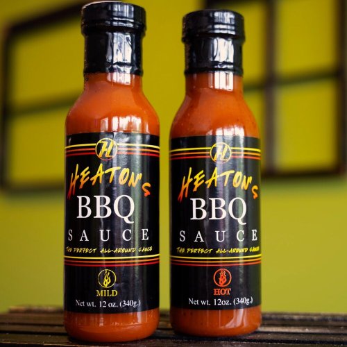 Two bottles of Heaton's BBQ sauce, labeled "Mild" and "Hot," on a colorful background. Each bottle is 12 oz.