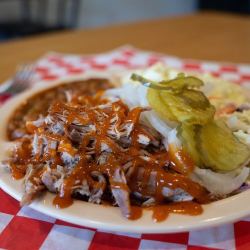 A plate of pulled pork topped with sauce, pickles, and sides of coleslaw and beans on a checkered tablecloth.