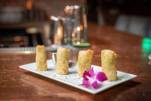 Four crispy spring rolls on a white plate, garnished with a purple flower and a small bowl of dipping sauce.