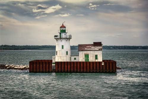 A white lighthouse with a green door stands on a rocky pier, surrounded by water under a cloudy sky.