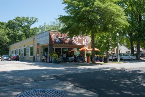 A cozy café with outdoor seating, surrounded by trees on a sunny day. Cars are parked nearby.