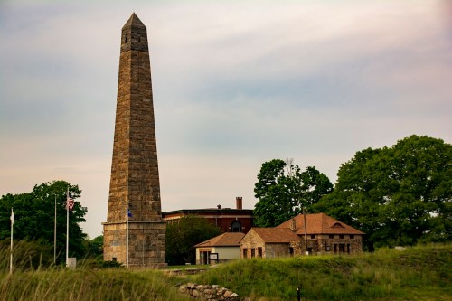 A tall stone obelisk stands beside a historic building, surrounded by greenery under a cloudy sky.